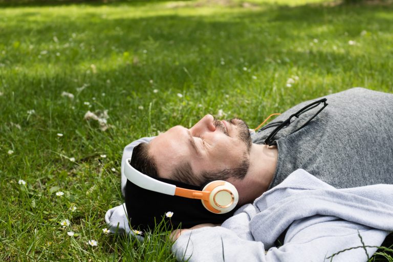 Relaxed man lying down on the grass and listening music with eyes closed.
